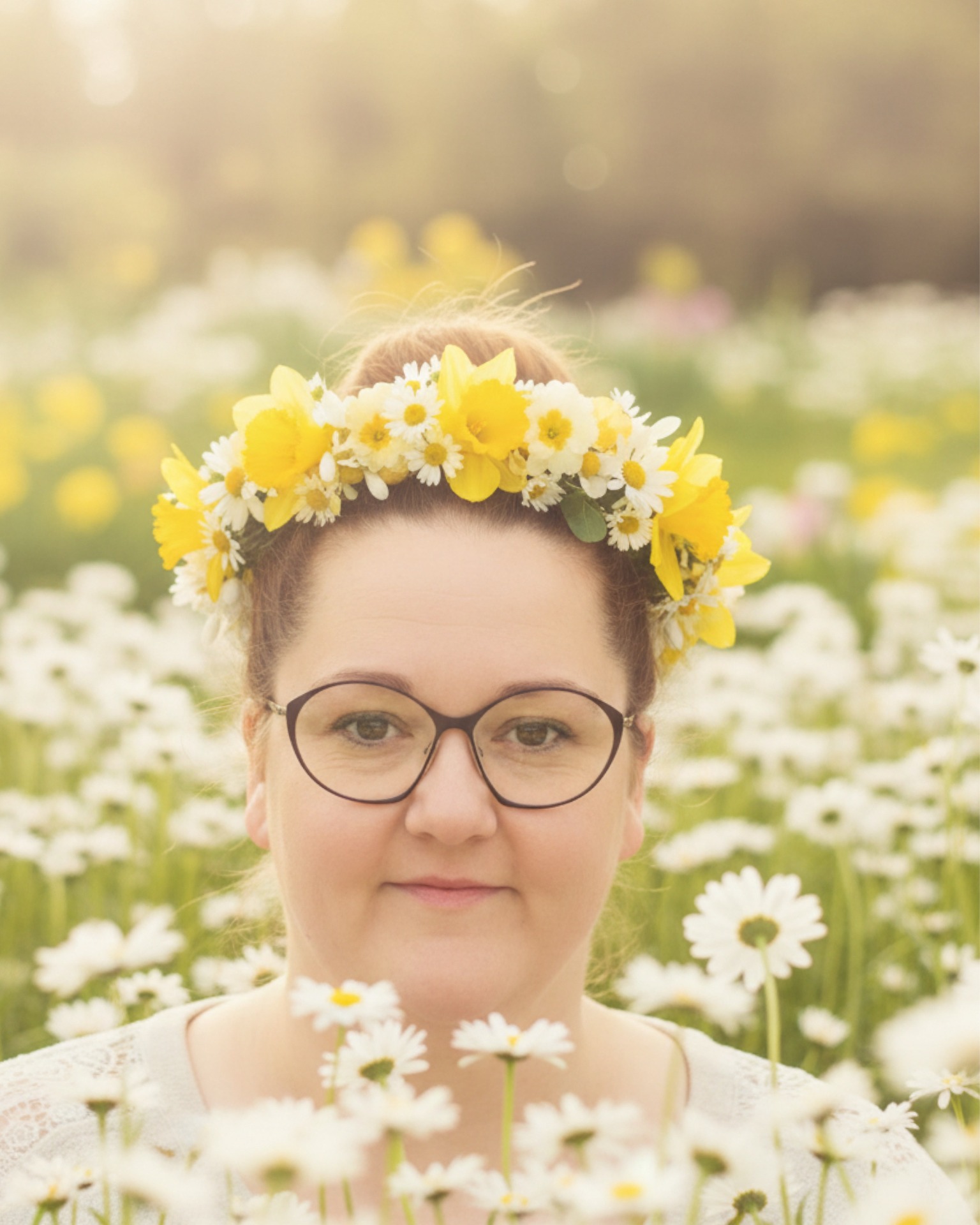 Porträt einer Frau mit Brille, Blumenkranz aus gelben und weißen Blüten im sonnigen Wiesenhintergrund.